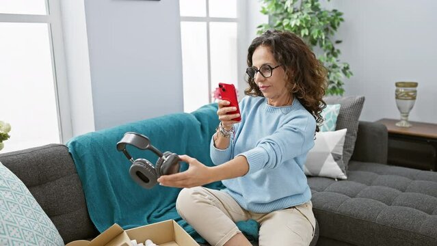 A middle-aged woman examines new headphones while holding a smartphone in a cozy living room setting.
