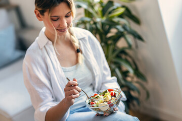 Young woman eating healthy salad while sitting on the kitchen at home.
