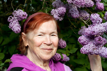 Outdoor close-up portrait of old woman. Beautiful elderly woman smiling against background of blooming lilacs in spring park.