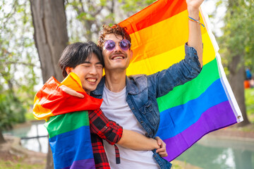 Gay couple celebrating love waving lgbt flag in a park - obrazy, fototapety, plakaty
