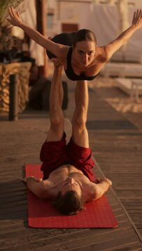 Vertical video: a guy and a girl doing a combination of acrobatics and yoga. The girl is in the flying position, and the guy is holding her with his legs on a sunny beach covered with boards. Acroyoga