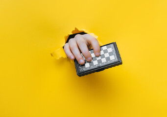 A child's hand emerges from a torn hole in yellow paper and holds a small chessboard. The concept of intellectual development and games that develop thinking.