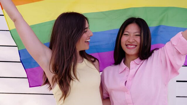 Young couple in love lesbian girls holding up rainbow flag outdoors. Two laughing multiracial cute women enjoying pride celebration together with white background. Friendship between female gay people
