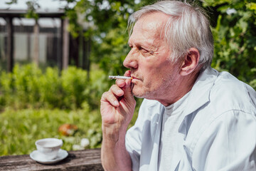 Elderly man smoking a cigarette, wearing a white shirt, outdoors. Reflective, personal moment,...