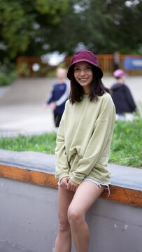 A brunette girl in a green sweater put one foot on a skateboard and sits against the backdrop of a skate park. Youth hobby, pastime, leisure