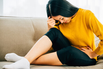 A woman of Asian descent suffers stomach ache on a living room couch at home. Depicting discomfort...