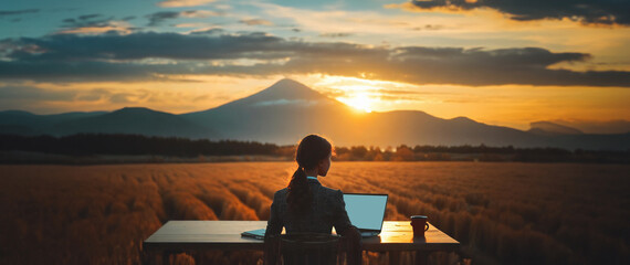 A panoramic, widescreen photo of a brown female office worker sitting at a desk overlooking a mountain range and a field at sunrise -  a laptop and a cup of coffee sit on the desk; working remotely