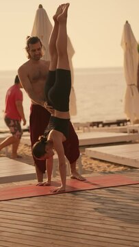 Vertical video: a brunette guy in red pants with a bare torso helps a girl in a black sports summer uniform to stand on her hands with her head down on a sunny beach on a red carpet