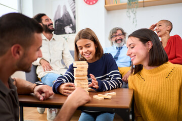 Happy diverse generation family gathered playing Jenga at home. Caucasian people together making domestic life with board games. Digital detox. Wooden block out of tower and others are watching