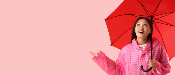 Beautiful young Asian woman in raincoat with umbrella pointing at something on pink background