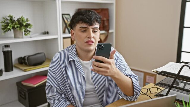A young man in a striped shirt ponders while holding a smartphone in a modern office setting.