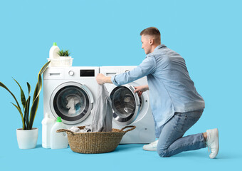 Handsome young man putting laundry into washing machine on blue background