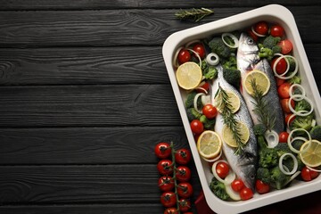 Raw fish with lemon in baking dish and vegetables on black wooden table, flat lay. Space for text