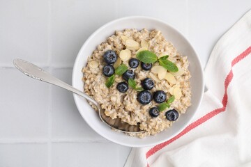 Tasty oatmeal with blueberries, mint and almond petals in bowl on white tiled table, top view