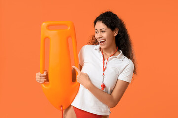 Beautiful young happy African-American female lifeguard pointing at rescue buoy on orange background