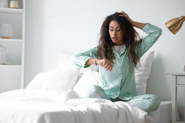 Overslept young African-American woman looking at wristwatch in bedroom
