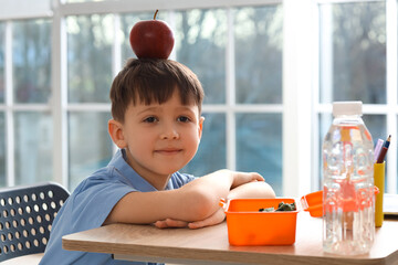 Cute little schoolboy with apple on his head during lunch in classroom