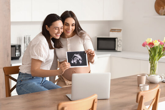 Young pregnant lesbian couple with sonogram image video chatting in kitchen