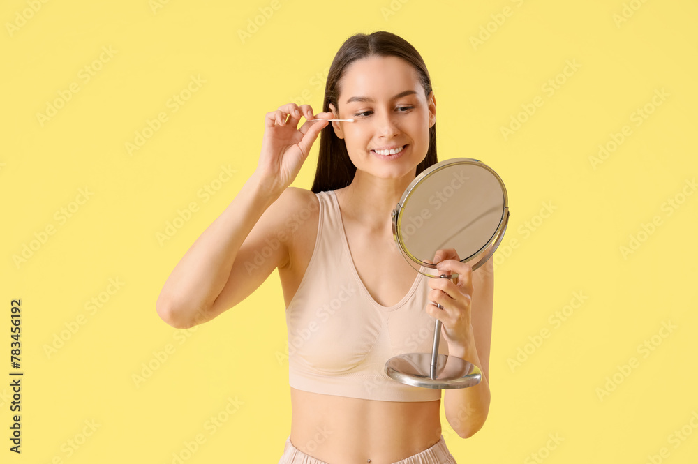 Sticker Young woman with cotton bud and mirror removing makeup on yellow background