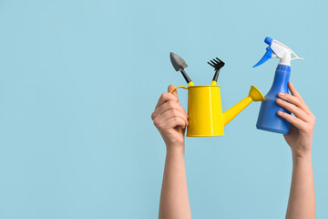 Female hands holding watering can with gardening rake, shovel and sprayer on blue background