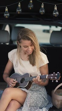 Vertical video: A blonde girl plays the ukulele in the trunk of a black car decorated with light bulbs