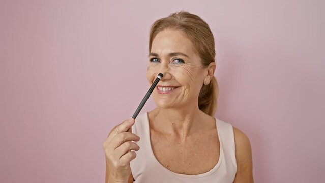 Mature caucasian woman applies makeup with a brush against a pink wall, embodying beauty and elegance in a serene setting.