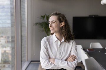 Pretty young 25s office employee, businesswoman posing in modern meeting room, smiling, looking out window to the big city view, full of aspirations. Career opportunities and professional achievements - obrazy, fototapety, plakaty