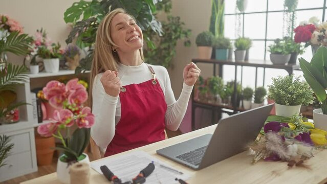Joyful young blonde florist woman sitting at flower shop table, victoriously screaming in happy excitement celebrating win, gesturing success!
