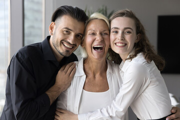 Three multigenerational colleagues relish success in office. Portrait of laughing attractive family hugging standing together in modern flat, feel love, enjoy carefree weekend at home. Unity, bonding - obrazy, fototapety, plakaty