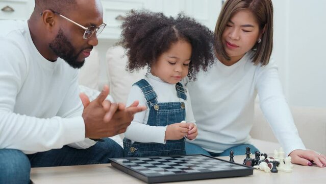 Little kid girl playing chess board at home. Adorable multiracial daughter in living room, enjoy playing piece of chess with parent. Joyful leisure childhood