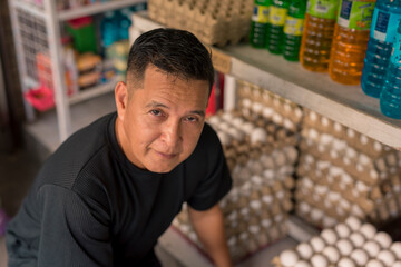 A middle-aged Asian man, an egg vendor, crouching in front of multiple trays of eggs at a small store, with a warm smile on his face. An egg trader or retailer.