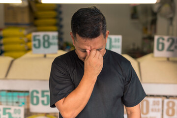 A owner of a rice trading business appears tense and worried, standing in front of his shop with...