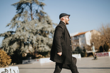 A fashionable man in winter apparel confidently strides forward in a sunlit urban park setting, showcasing a casual yet sophisticated style.