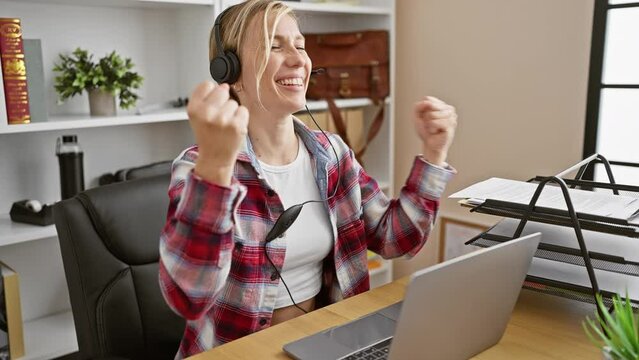 Excited young blonde business woman celebrates victory with raised arms in carpentry workshop, beaming with success while using her laptop and headphones