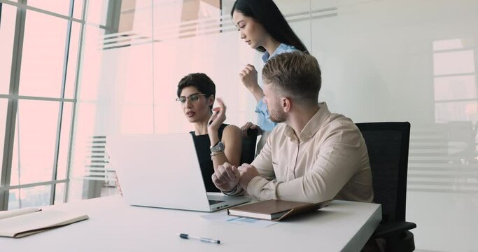 Serious multiethnic business team of three discussing online content on laptop, pointing at monitor, speaking, testing new job application, watching project presentation on Internet