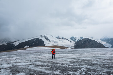 Man on ice pass with view to big glacier tongue among sharp rocks and large snow-capped mountains in rainy low clouds. Guy against dark atmospheric mountain silhouettes in rain in gray cloudy sky. - 783493900
