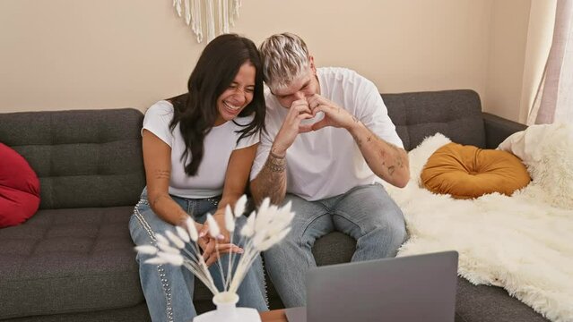 A joyful man and woman making a heart shape with hands while having a video call in a cozy living room.