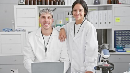 A smiling man and woman in white lab coats stand together in a modern laboratory, portraying teamwork and scientific research.