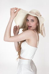 Portrait of a young blonde woman with an open mouth looking over shoulder on a white background. She is wearing a white corset top and pants and her hands are raised, holding the brim of a straw hat