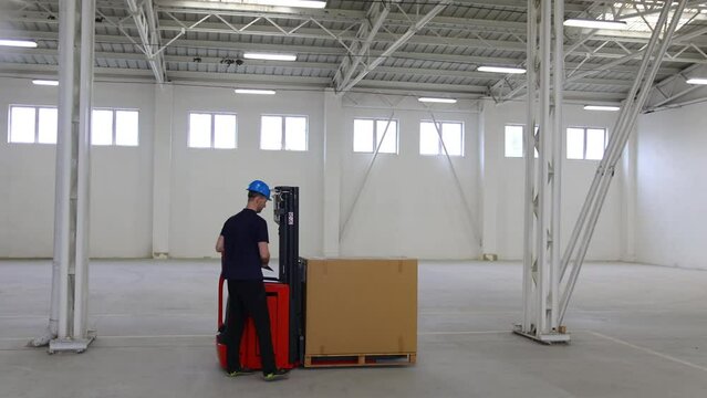 A young man in a helmet, a worker in a warehouse, checking and transporting pallet in metal hall. A warehouse of packages of products, the concept of transportation