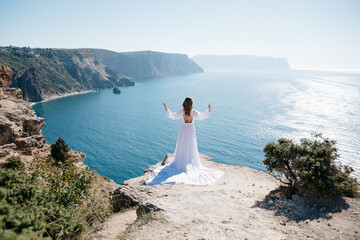 A woman stands on a cliff overlooking the ocean in a white dress. She is looking out at the water and she is in a state of peace and serenity. Concept of calm and tranquility.