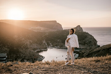 A woman stands on a hill overlooking a body of water. The sun is setting in the background, casting a warm glow over the scene. The woman is enjoying the view and taking in the beauty of the landscape