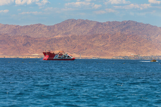 Red and white modern boat sailing on a tranquil blue sea. The backdrop features rugged mountains under a clear sky. The mountains, though barren, add a majestic element to the scene. Red sea, Israel