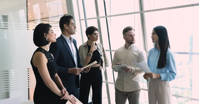 Multiethnic business team of serious coworkers standing at office large window for briefing, talking, discussing work tasks, teamwork strategy, listening to male Indian boss