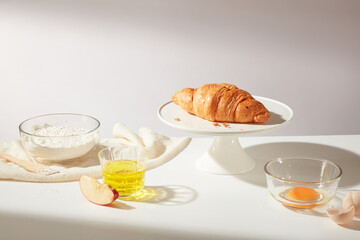 White background with a delicious croissant featured on a round white dish decorated by some ingredients for baking such as oil, egg, flour surrounded. Copy space photo for bakery