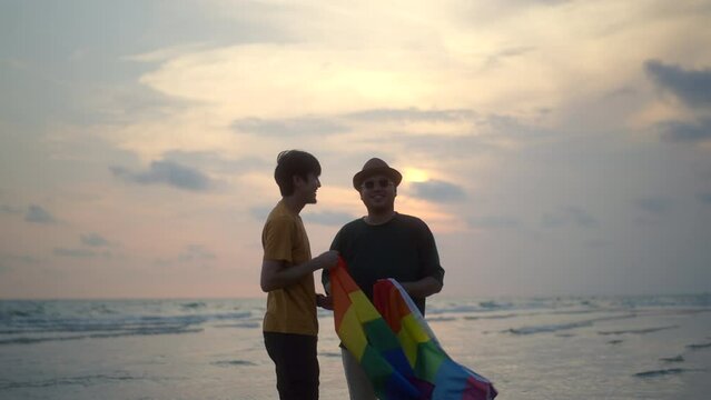 Romantic moment of bisexual gay male couple walking on the beach with their arms crossed with rainbow flag. LGBTQ man couple have a romantic moment on the beach. LGBT and homosexual concept.