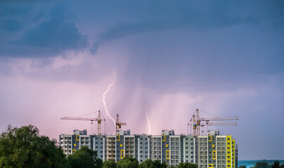 An apartment building with construction cranes against the background of the night sky where you can see lightning