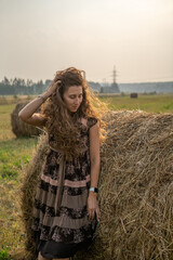 a beautiful girl with curly hair rests on a field next to a haystack on a warm and sunny summer day