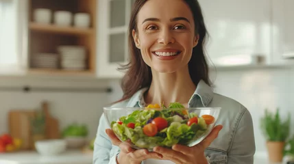 Fotobehang A happy young beautiful woman or girl holding a bowl of fresh organic green vegetable salad in a light kitchen. Eating healthy food. Concept of Diet, Fitness, Healthy lifestyle, vegetarianism.  © Lahiru