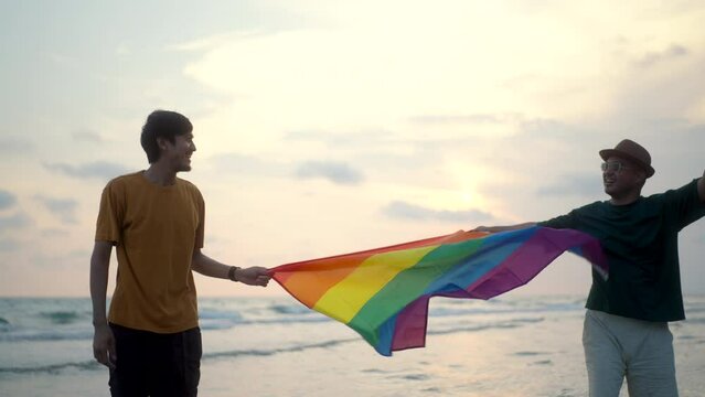 Romantic moment of bisexual gay male couple walking on the beach with their arms crossed with rainbow flag. LGBTQ man couple have a romantic moment on the beach. LGBT and homosexual concept.